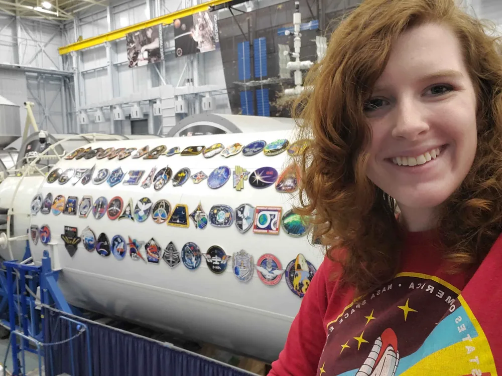 Haleigh beside an ISS module mockup and mission-patch wall at a NASA facility